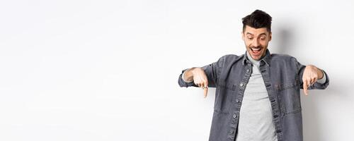 Excited european man looking and pointing fingers down, looking happy at advertisement, standing in denim jacket on white background photo