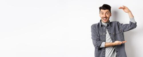 Cheerful guy smiling and showing big size, showing large thing and looking excited, standing against white background photo