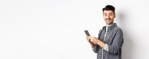 Handsome young man using smartphone and looking at camera. Caucasian guy chatting on phone with happy face, standing on white background photo
