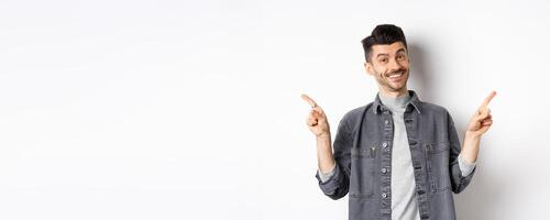 Young handsome guy with moustache pointing fingers sideways, showing two logos, smiling happy at camera, standing in casual clothes on white background photo