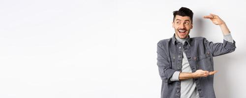 Excited guy looking at logo and showing big size thing, shaping large object and smiling amazed, standing in denim jacket on white background photo