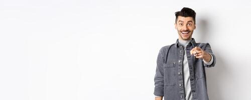 It is you. Excited smiling man pointing fingers at camera with cheerful face, recognize person, standing in denim jacket on white background photo