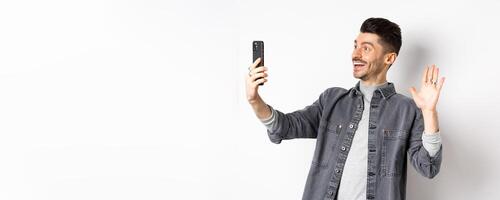 Young man chatting on mobile phone, waiving hand at smartphone camera, greeting friend online, standing on white background photo