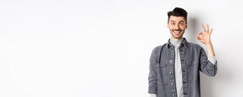 Smiling handsome man showing OK sign, like good product, recommending advertisement, standing satisfied on white background photo