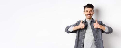 Handsome smiling man showing thumbs-up in approval, praise good work, satisfied with nice choice, recommending something cool, standing on white background photo