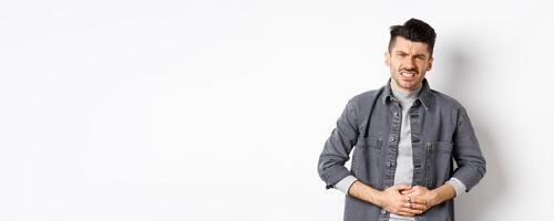 Man with stomach ache frowning and touching belly, having food poison, suffer diarrhea, standing on white background photo