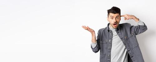 Are you crazy. Angry man scolding person for stupid actions, pointing at head temple and raising hand confused, complaining while standing on white background photo