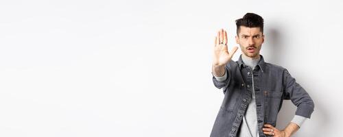 Serious man tell to stop, prohibit something and frowning, say no, standing displeased with stretch out hand against white background photo