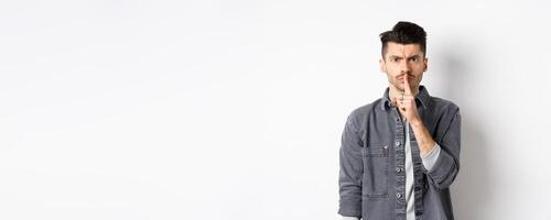 Be quiet. Angry man frowning and shushing, telling to calm down, press finger to lips as scolding bad behaviour, standing on white background photo