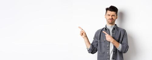 Skeptical and disappointed young man pointing fingers left, grimacing and shaking head in rejection, dislike bad product, standing on white background photo