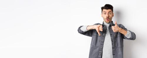 Excited guy show thumbs up and down judging and making decision, rate something interesting, standing on white background photo