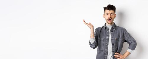 Confused and annoyed guy raising hand up and complaining, asking wtf happened, cant understand, standing on white background photo