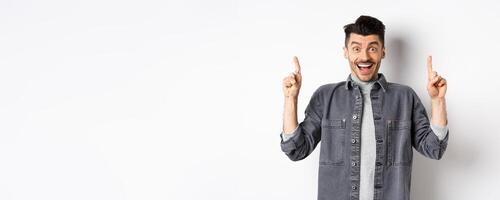 Excited handsome guy pointing fingers up, smiling happy and showing special deal, standing on white background photo