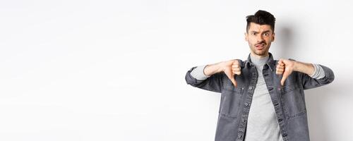 Disappointed guy showing thumbs down and frowning, looking skeptical at something bad, dislike and disapprove awful thing, standing on white background photo