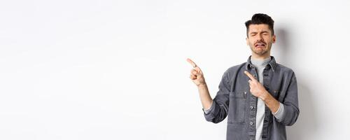 Sad crying man pointing fingers left at logo, sobbing and whining, being jealous or upset, standing on white background photo