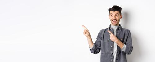 Handsome smiling guy checking out advertisement, looking and pointing left with excited face, standing on white background photo