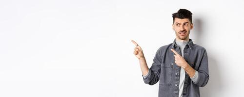 Skeptical and doubtful young man pointing fingers left at logo, look with dislike and confusion, standing on white background photo