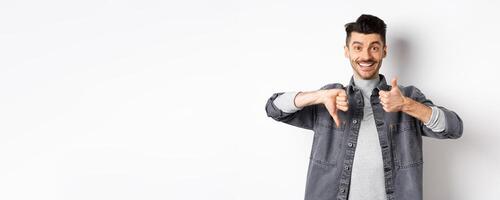Happy smiling guy showing average gesture, thumbs down up, cant decide, judging something medium, standing on white background photo