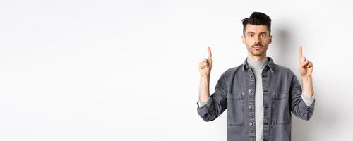 Serious caucasian guy with moustache, pointing fingers up at empty space and look at camera, white background photo