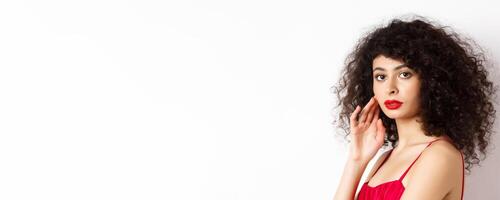 Close up of elegant woman with curly hair and red lips, looking at camera and touching face, standing in red dress on white background photo