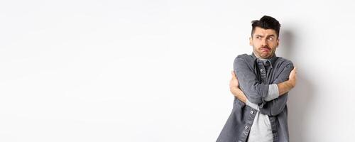 Displeased young man feeling discomfort, hugging himself and staring left at something embarrassing or exposing, standing uncomfortable on white background photo