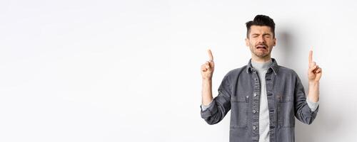 Sad and miserable guy crying while pointing fingers up, showing disappointed promo, sobbing and whining, standing on white background photo