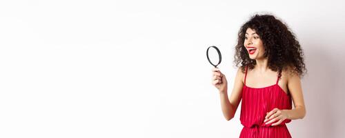 Excited woman in red dress look through magnifying glass and smiling, found interesting promo, standing on white background photo