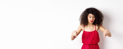Intrigued beautiful woman with red lips and dress, pointing and looking down at banner, reading logo, standing over white background photo