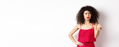 Confused woman with curly hair, wearing red dress, frowning and shaking hand, cant understand something, looking hesitant, standing on white background photo