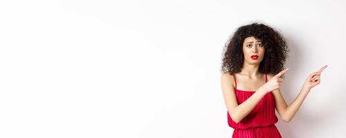 Confused and sad woman with curly hair, wearing evening dress and makeup, pointing fingers right and frowning upset, standing on white background photo