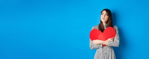 Valentines day. Sad heartbroken girl sobbing, looking aside at empty space and crying upset, hugging big red heart, feeling lonely without lover, standing alone on blue background photo