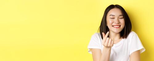 Valentines day and women concept. Close up of pretty asian girl in white t-shirt, smiling and showing finger heart, I love you gesture, standing over yellow background photo