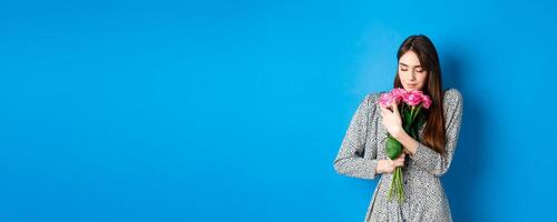 Valentines day concept. Passionate and romantic young woman hugging bouquet of gift roses, smelling flowers from lover, standing on blue background photo