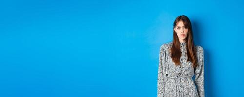 Moody frowning girl in dress looking at unfair thing, standing offended or upset against blue background photo