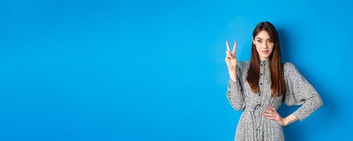 Young beautiful girl in dress with natural long hair, showing number two with fingers and smiling, standing against blue background photo