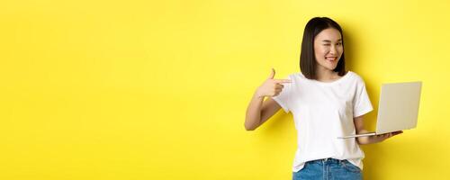 E-commerce and shopping concept. Good-looking asian girl winking at camera, smiling and pointing finger a laptop, showing something, standing over yellow background photo