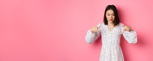 Concerned asian woman peeking down and pointing hand at disturbing banner, frowning upset, standing over pink background photo