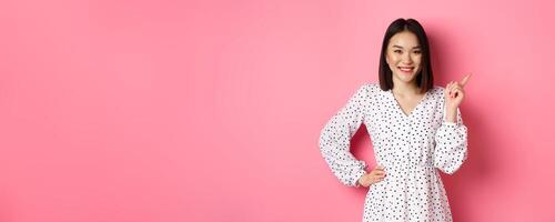 Beautiful asian female model smiling, pointing finger at upper right corner copy space, showing advertisement banner, standing over pink background photo
