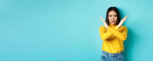 Serious and assertive korean woman showing cross stop gesture, frowning and telling no, prohibit action, disapprove something bad, standing over blue background photo