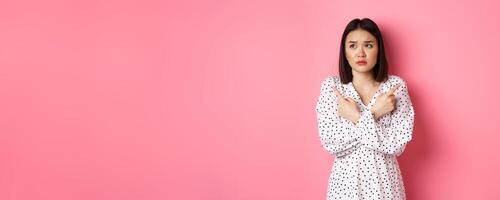 Concerned asian woman having doubts, pointing sideways and looking left with hesitant and sad face, need help with choice, standing over pink background photo