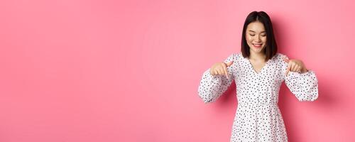 Cute romantic asian woman smiling, pointing fingers and looking down with pleased face, showing product discount, standing over pink background photo