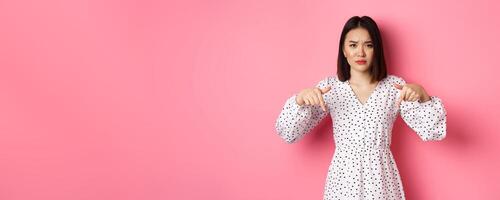 Disappointed asian female model pointing fingers down at promo offer, frowning and staring at camera upset, standing over pink background photo