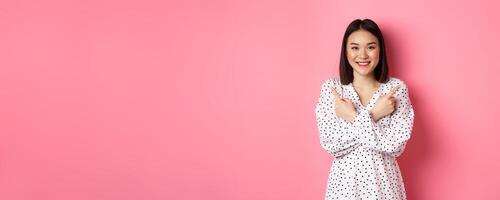 Beautiful asian girl in romantic dress pointing fingers sideways, showing two variants on shopping, smiling at camera, standing against pink background photo