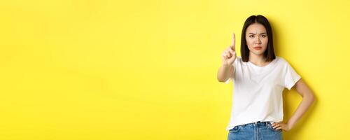 Confident and serious woman tell no, showing extended finger to stop and prohibit something bad, frowning and looking at camera self-assured, standing over yellow background photo