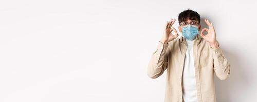 Covid-19, health and real people concept. Excited and impressed guy in sterile face mask showing OK signs in approval, praise cool thing, standing amused on white background photo