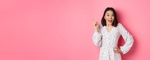 Cute asian woman in dress pointing upper left corner copy space. Korean model showing advertisement, standing over pink background photo