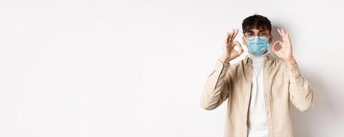 Covid-19, health and real people concept. Handsome guy in glasses and medical mask showing okay gesture in approval, recommend use preventive measures from corona, white background photo