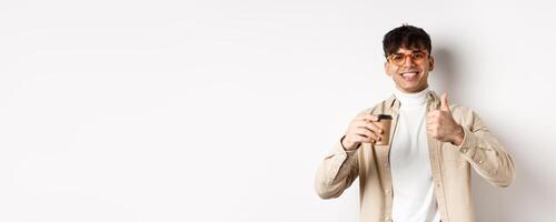 Real people. Cheerful smiling guy in glasses drinking coffee and showing thumb up, recommending cafe, standing on white background photo
