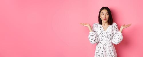 Confused and clueless asian girl shrugging, staring at camera puzzled, dont know what to do, spread hands sideways, standing over pink background photo