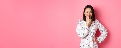 Beautiful Korean woman in trendy dress asking to keep secret, hushing with soft smile, planning a surprise, standing over pink background photo
